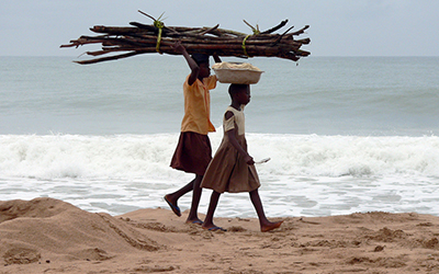 Young Ghanian children walk across beach carrying things on their heads