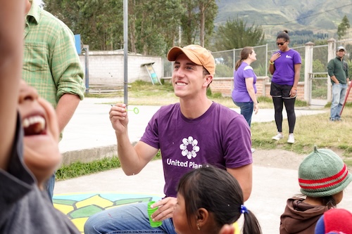 Young man kneeling down with a hat on in a purple shirt that has the United Planet logo. Around him are children playing and two other volunteers.